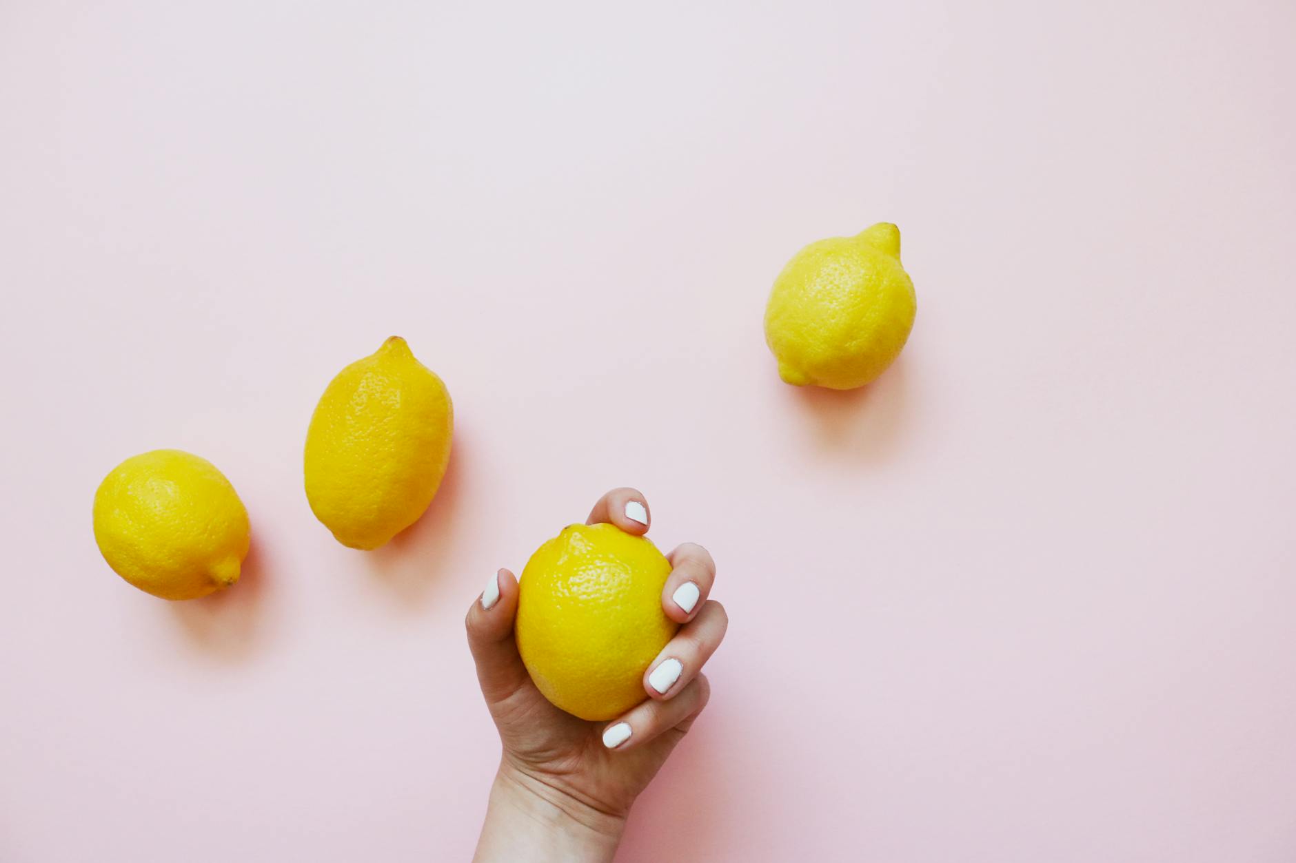Hand with white nails holding a fresh lemon on a soft pink background, surrounded by additional lemons